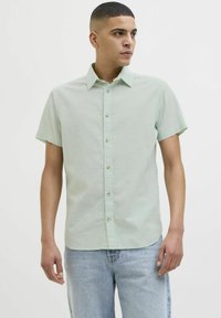 Camisa de manga corta verde claro con diseño de botoneo, que presenta un cuello clásico y una textura sutil, combinada con pantalones de mezclilla azul claro.