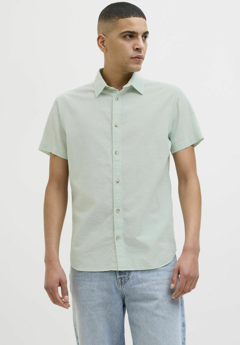 Camisa de manga corta verde claro con diseño de botoneo, que presenta un cuello clásico y una textura sutil, combinada con pantalones de mezclilla azul claro.