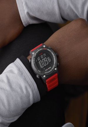Digital sports watch with red strap on wrist, showing time and date, worn over black and white long-sleeve clothing.