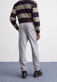 En person sedd bakifrån bär en randig svart och grön långärmad tröja och grå sweatpants, i kombination med svarta skor, stående på en matta.