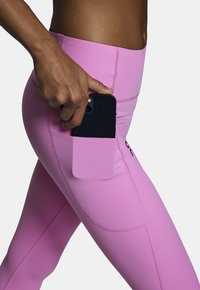 Różowe legginsy z boczną kieszenią, w której trzymany jest czarny smartfon. Wykonane z elastycznego materiału, charakteryzujące się płaskimi szwami i gładką fakturą.