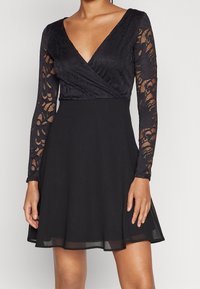 Svart klänning med genomskinliga spetslånga ärmar, djup V-ringning och omlottliv, samt A-linjeskirt, som bärs av en stående person, beskuren vid nacken och mitt på låret.