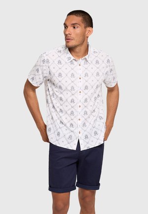 Hombre con el pelo corto que lleva una camisa blanca de manga corta con botones y estampado, y unos pantalones cortos azul marino remangados, mirando hacia la izquierda.