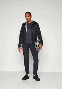 Nő fekete dzsekiben, sötét nadrágban, szürke kerékpáros sisakot tartva, egy világos háttér előtt áll, laza testtartással a kamerába néz.