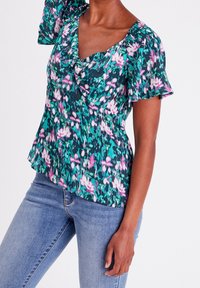 Květovaný peplum top v zelené barvě s růžovými a bílými vzory, který má krátké volánkové rukávy a kulatý výstřih, vyrobený z lehké tkaniny.