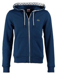 Mörkblå zip-up hoodie med köttpåse, vita dragsko och geometriskt mönster på insidan av huvan. Har en liten logotyp på bröstet.