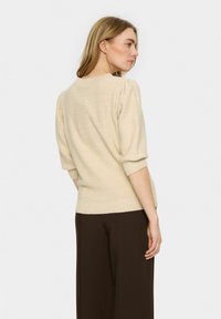 Beiza adīta džemperis ar puffed piedurknēm, ribotām aprocēm un apakšu; valkāts ar brūnām platām biksēm; gluda tekstūra bez redzamām rakstu kļūdām.