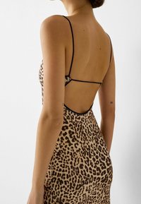 Žena v šatech s leopardím potiskem bez zad a tenkými černými ramínky, ukazující tetování na obou horních pažích na jednobarevném bílém pozadí.