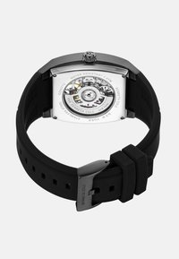 Quadratisches Gehäuse aus Edelstahl mit transparentem Gehäuseboden, schwarzem Gummiband und silbernem Zubehör. Wasserfest bis 3 ATM.