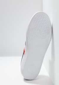 La suola della sneaker bianca presenta una gomma texture con un motivo geometrico. La parte superiore ha accenti scuri e un dettaglio del logo rosso.