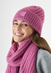 Roze gebreide muts met ribbelstructuur en een logopatch, gecombineerd met een bijpassende roze chunky sjaal, gestyled met een grijze jas.