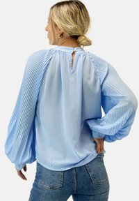Lichtblauwe blouse met textuurplooien op de mouwen, een keyhole achterkant met een knoopsluiting en een relaxed fit. De stof is zacht en vloeiend.