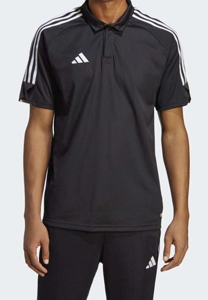 Homme portant un polo de sport Adidas noir et un pantalon avec des bandes blanches sur les épaules et des logos Adidas sur la poitrine et la cuisse.