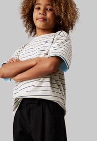 T-shirt a righe con maniche corte in crema e blu, con polsini blu e un piccolo logo ricamato. Abbinato a pantaloni neri.
