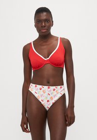 Top de biquíni vermelho canelado com acabamento em branco, combinado com parte de baixo de cintura alta com um padrão colorido de frutas sobre um fundo branco.