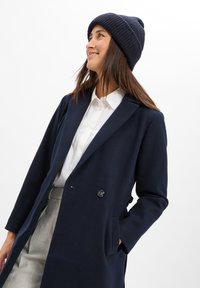 Navyblauer Wollmischungsmantel mit modernem Schnitt, doppelt geknöpftem Vorderteil, eingeknöpften Revers und seitlichen Taschen. Kombiniert mit einer gerippten navyblauen Mütze.