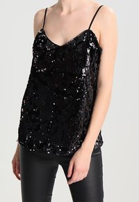 Haut noir sans manches en tissu pailleté, présentant un col en V et une coupe ample. La texture est brillante, ajoutant un intérêt visuel.