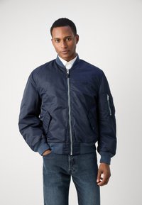 Marine bomberjakke med glidelås foran, ribbestrikkede mansjetter og liv. Har sidelommer, en glidelåslomme på ermet og glatt tekstur.