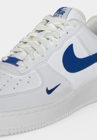 Άσπρο παπούτσι Nike Air Force 1 με μπλε λογότυπα Swoosh, λευκά κορδόνια, διάτρητη μύτη και ετικέτα λογότυπου στο κάτω κορδόνι.