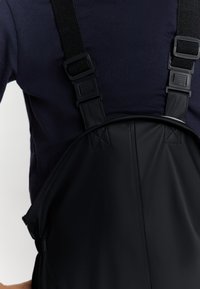 Juodas vandeniui atsparus priekalnis su reguliuojamomis dirželių ir plastikinėmis sagtimis. Lygus paviršius su subtiliu blizgesiu. Yra įstrižų siūlių detalė.