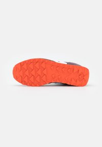 Oranssi kuminen lenkkarin ulkopohja, jossa on kolmionmuotoiset urat pitävyyttä varten ja teksturoitu pinta. Brändäys näkyvissä.