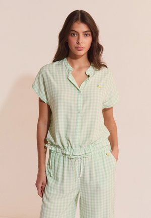 Junge Frau trägt eine hellgrün-weiß kariertes kurzärmeliges Bluse und passende Hosen mit Kordelzug in der Taille, steht vor einfachem Hintergrund.