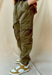 Olive green cargo pants in a lightweight fabric, featuring multiple pockets and elastic cuffs, paired with white and mustard sneakers.
