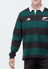 Persoon die een rugbyshirt met lange mouwen draagt, groen en zwart gestreept, met een witte kraag en All Blacks- en Adidas-logo's op de borst.