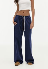 Kvinde iført marineblå vidde sweatpants med snøre, hvid cropped tanktop og brune ruskinds slip-on sko.