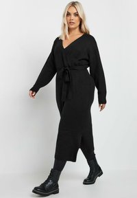 Musta kietaisumekko pehmeästä neulemateriaalista, jossa on syvä V-kaula-aukko, pitkät hihat, vyötäröllä solmittava vyö ja sivuhalkio.