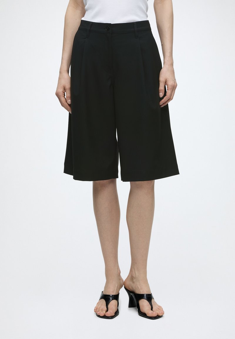 Femme portant un short noir ample mi-cuisses, un haut blanc et des sandales à talons noires ouvertes à l’avant, debout devant un fond uni.