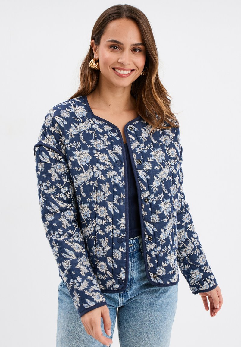 Veste matelassée bleu marine avec un motif floral blanc, col rond et fermetures à pression à l'avant. Manches longues et détail de tissu textur é.