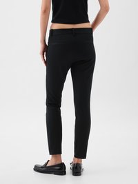 Nő slim-fit fekete nadrágban és fekete loferben áll, kezei lazán az oldalán, háttal a kamerának egy egyszínű háttér előtt.