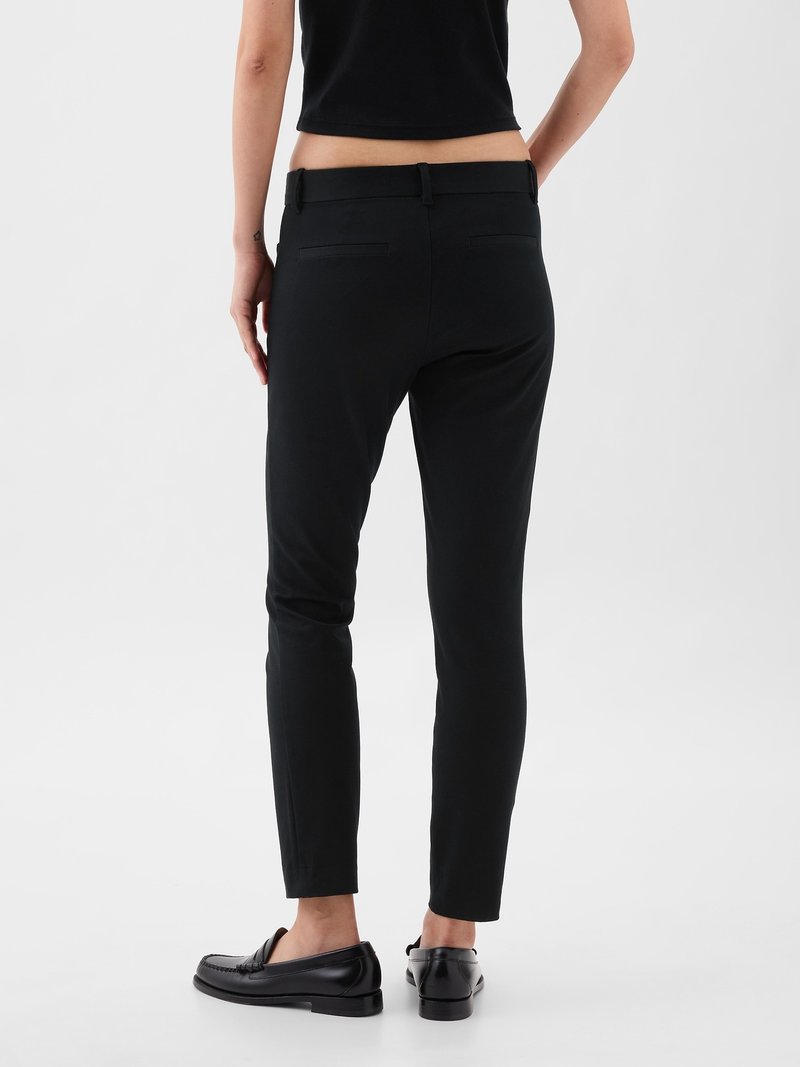 Femme portant un pantalon noir slim et des mocassins noirs, debout avec les mains relâchées sur les côtés, de dos face à l'appareil photo sur fond uni.