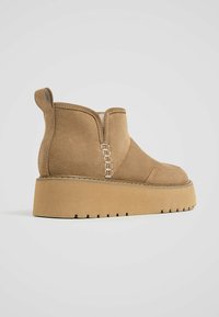 Bottines en daim marron avec des détails de couture sur le côté, un bout arrondi et une semelle épaisse et texturée. Un design simple et élégant.