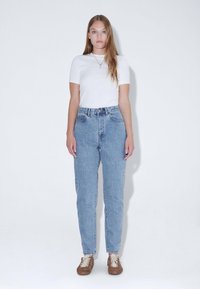 Světle modré džíny s vysokým pasem a rovnými nohavicemi, doplněné o přiléhavé bílé tričko a hnědé tenisky na tkaničky. Jednoduchý design, bez vzorů.