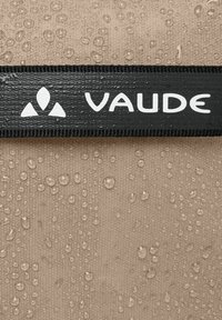 Logo VAUDE nero su materiale beige testurizzato con gocce d'acqua, evidenziando la sua caratteristica impermeabile e i dettagli del tessuto.