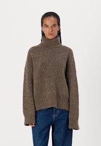Maglione marrone con texture, collo alto, spalle abbassate e maniche lunghe. Dettaglio orlo sfilacciato e vestibilità rilassata. Indossato con jeans blu.