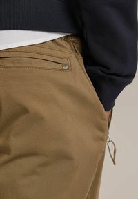Spodnie khaki wykonane z bawełny, z boczną kieszenią z metalowym okuciem oraz elastycznym paskiem w talii. Teksturowany materiał z widocznym szwem.