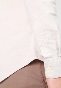 Camisa beige claro abotonada con mangas largas, con una textura lisa y un dobladillo curvo, que presenta dos botones pequeños y redondos en los puños.