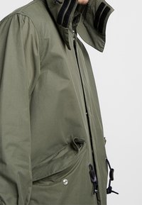Giacca verde oliva con zip, caratterizzata da un alto colletto con cinturini in velcro neri, cordini regolabili e due tasche frontali.
