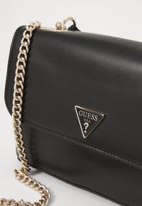 Černá umělá kožená crossbody taška se zlatým řetízkovým popruhem a trojúhelníkovým logem. Hladký povrch s pevnou strukturou.