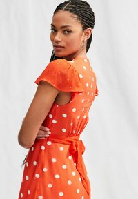 Abito rosso con pois bianchi, maniche svolazzanti corte, cinture in vita e un corpetto aderente. Tessuto liscio con una leggera lucentezza.