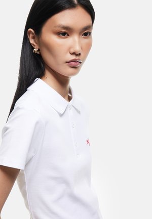 Młoda kobieta z długimi czarnymi włosami nosi złote kolczyki-koła i białą koszulkę polo z kołnierzykiem na jednolitym jasnym tle.