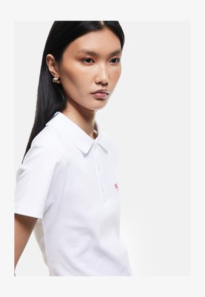 Joven mujer con cabello largo y negro lleva pendientes de aro dorados y una camiseta polo blanca con cuello contra un fondo claro y liso.