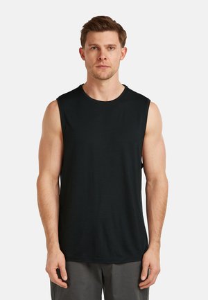 Mann mit kurzen braunen Haaren, der ein schlichtes schwarzes ärmelloses Shirt und graue Hosen trägt, vor einem weißen Hintergrund stehend.