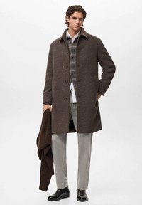 Abrigo de cuadros marrón con un diseño estructurado, cierre de botones y tejido suave. Combinado con un suéter gris, jeans claros y botas negras.