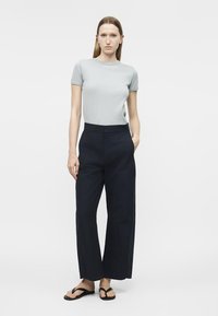 Femme debout, main dans la poche, portant un haut gris clair à manches courtes, un pantalon large taille haute foncé, et des sandales tongs noires.