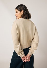Pull beige en maille avec un design court, ourlet côtelé et manches longues bouffantes. Porté avec un jean denim foncé. Texture douce et coupe décontractée.