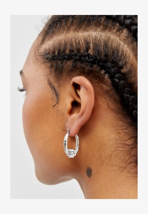 Srebrna kolczyk w kształcie obręczy z grawerowanymi detalami i teksturowanym wzorem, noszony na warkoczu. Zbliżenie na kolczyk i ucho.